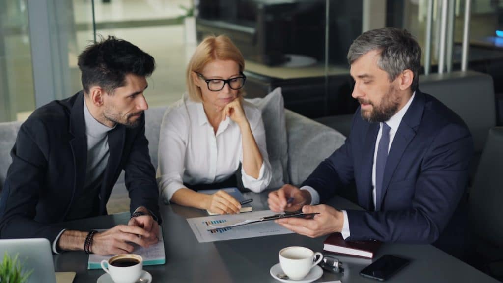 Three professionals in a modern office discuss documents and a tablet around a conference table, coffee cups nearby.