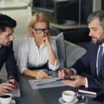 Three professionals in a modern office discuss documents and a tablet around a conference table, coffee cups nearby.