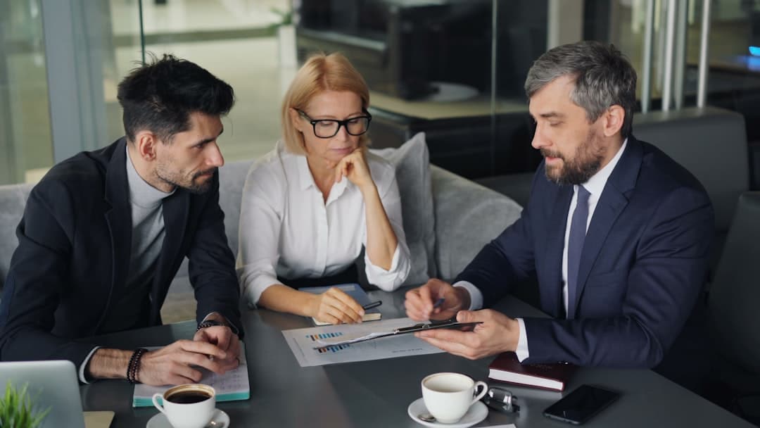 Three professionals in a modern office discuss documents and a tablet around a conference table, coffee cups nearby.