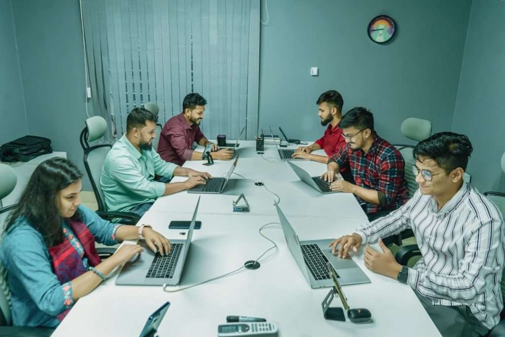Group of six colleagues around a white conference table, each with a laptop, collaborating in a modern office setting.
