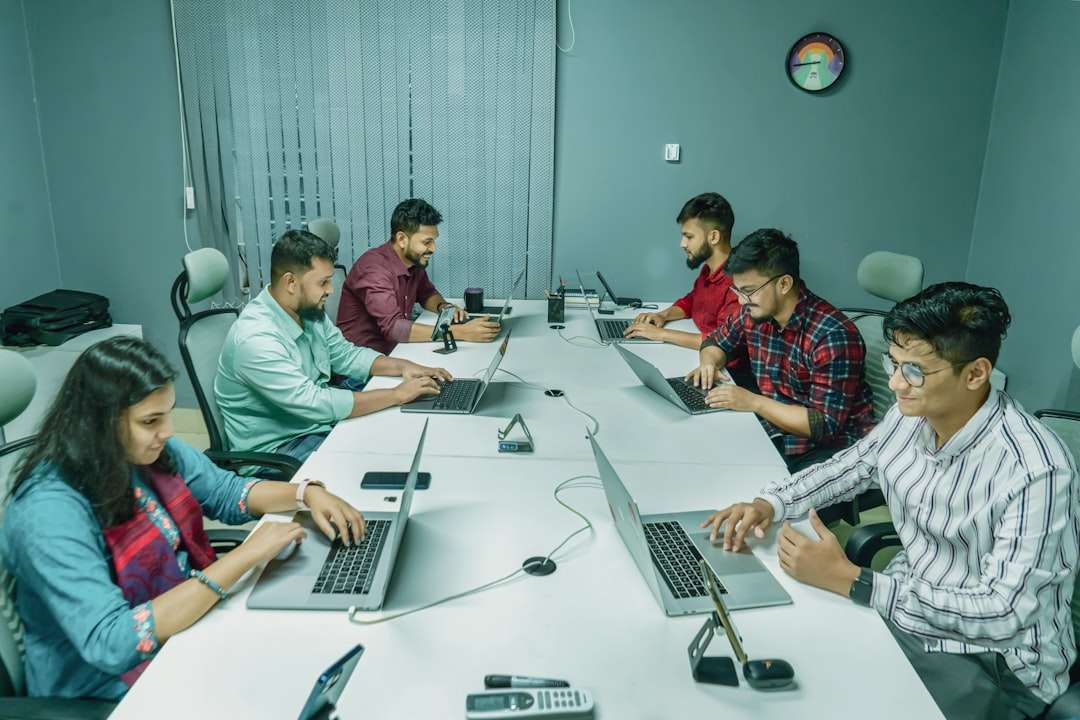 Group of six colleagues around a white conference table, each with a laptop, collaborating in a modern office setting.