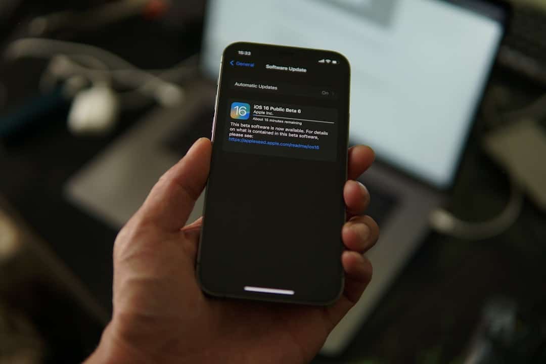 Hand holding an iPhone showing the Software Update screen for iOS 16 Public Beta 6 with 'Automatic Updates' option visible