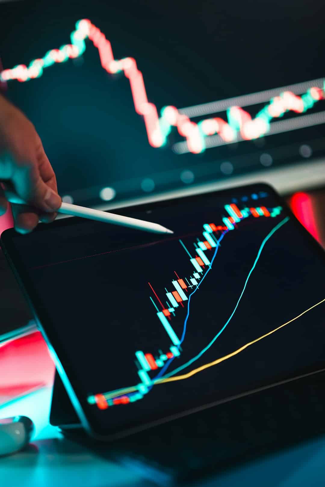 Trader's hand with a stylus on a tablet displaying colorful candlestick stock charts and moving averages.