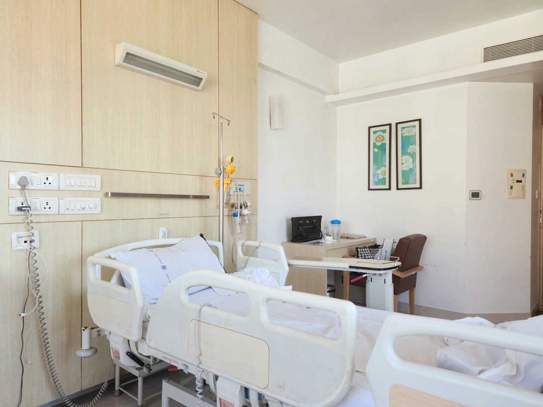 Hospital patient room with a bed, IV pole, and medical equipment along a wood-paneled wall; a desk and chair are in the background with two framed daisies pictures on the wall