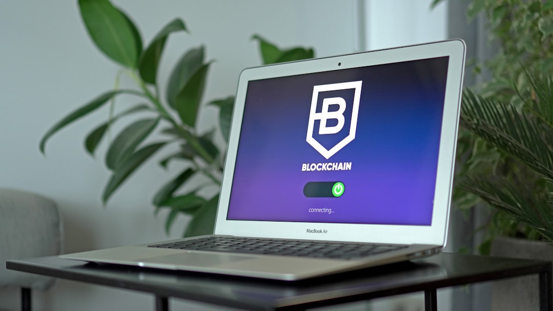MacBook Air on a dark table showing a blockchain app loading screen with a white shield logo and 'BLOCKCHAIN' text.