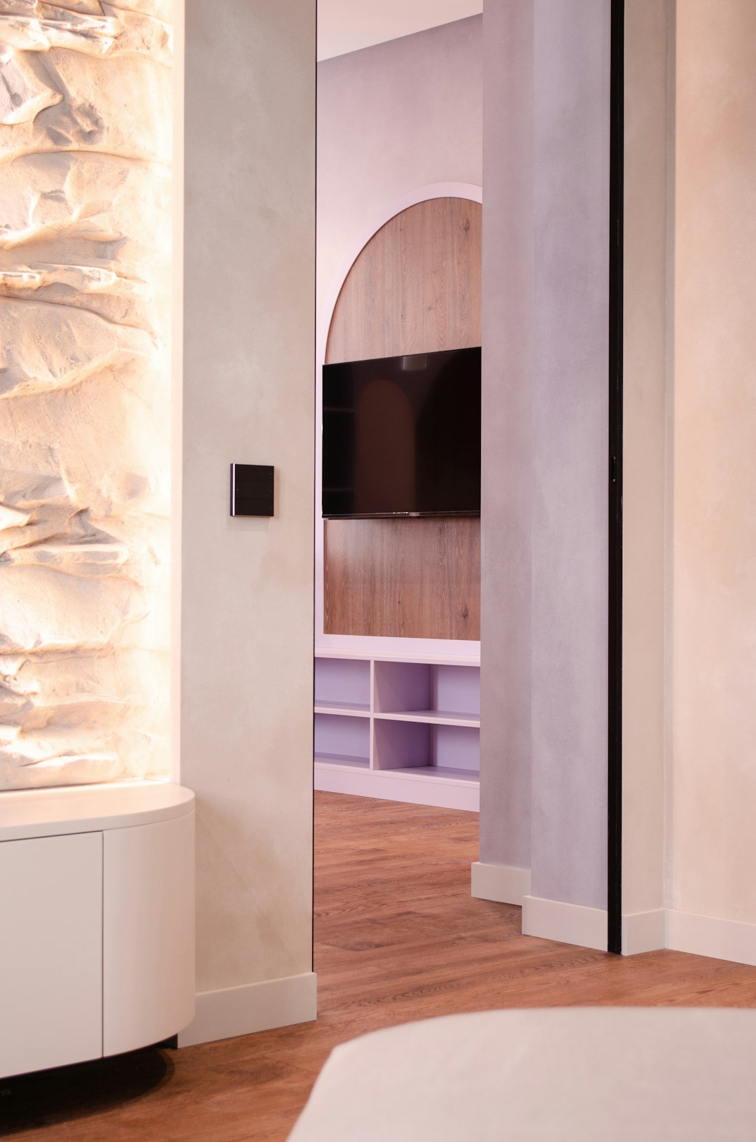 Doorway opening into a modern living space with a wall-mounted TV on wood paneling and a white cubby shelf beneath; stone wall to the left.