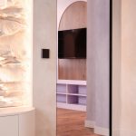 Modern hallway into a living area with a wall-mounted TV in a wooden panel niche and open cubby shelves beneath, flanked by light gray walls and a stone feature on the left.