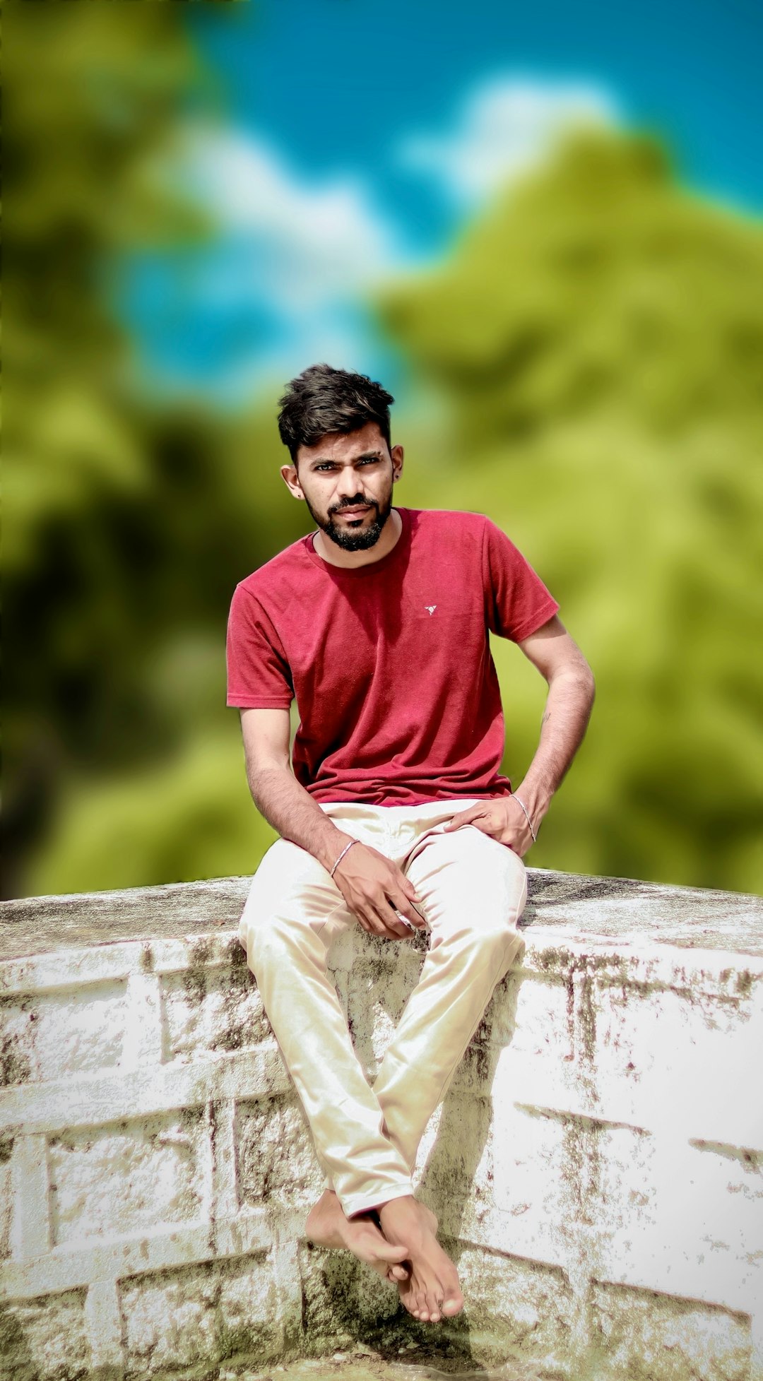 Young man in a red t-shirt sits on a weathered white concrete wall outdoors, barefoot and relaxed.