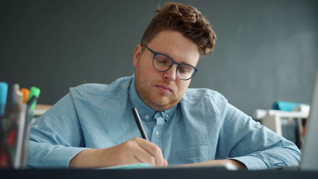 Man with blue shirt and glasses writing with a pen at a desk, concentrating on his work in a classroom or office setting.