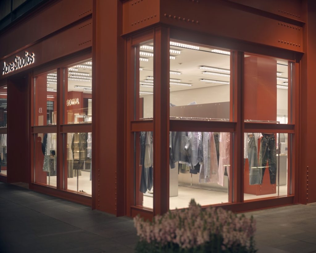 Acne Studios storefront at dusk with large glass windows showing clothing on display inside a red exterior storefront