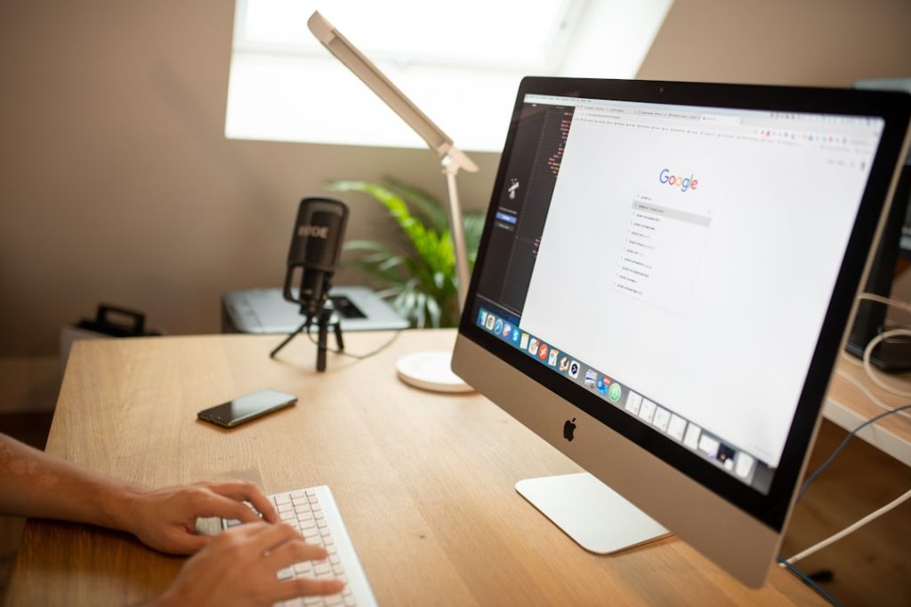 Person typing on a white keyboard at a wooden desk with an iMac, a microphone on a stand, a smartphone, a desk lamp, and a plant in the background.