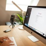 Person typing on a white keyboard at a wooden desk with an iMac, a microphone on a stand, a smartphone, a desk lamp, and a plant in the background.