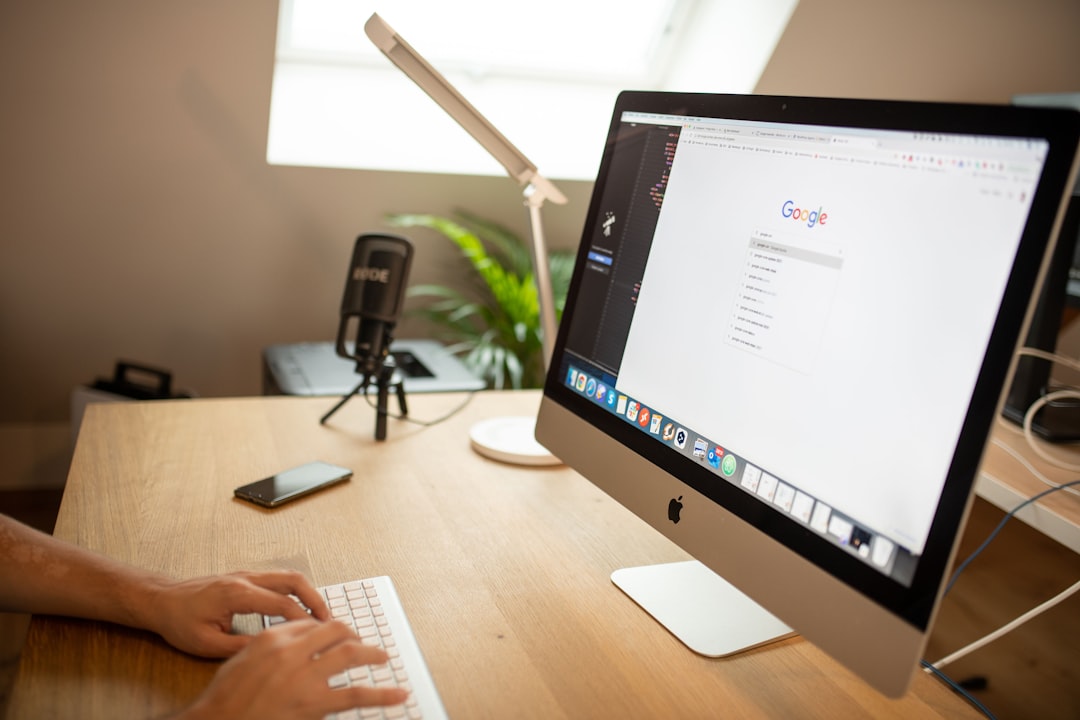 Person typing on a white keyboard at a wooden desk with an iMac, a microphone on a stand, a smartphone, a desk lamp, and a plant in the background.