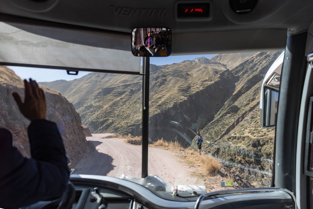 View from inside a bus as it travels a dusty mountain road; a hiker stands on the roadside ahead near a steep canyon.