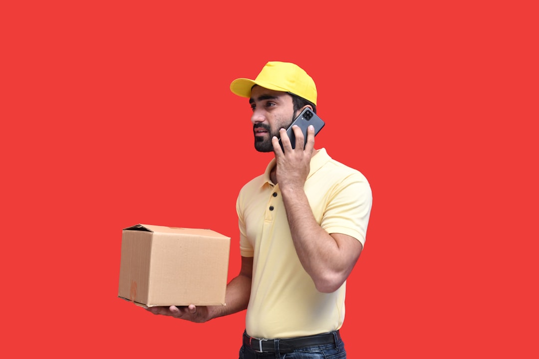 Delivery worker on a call, holding a cardboard box, wearing a pale yellow shirt and cap on a red background.