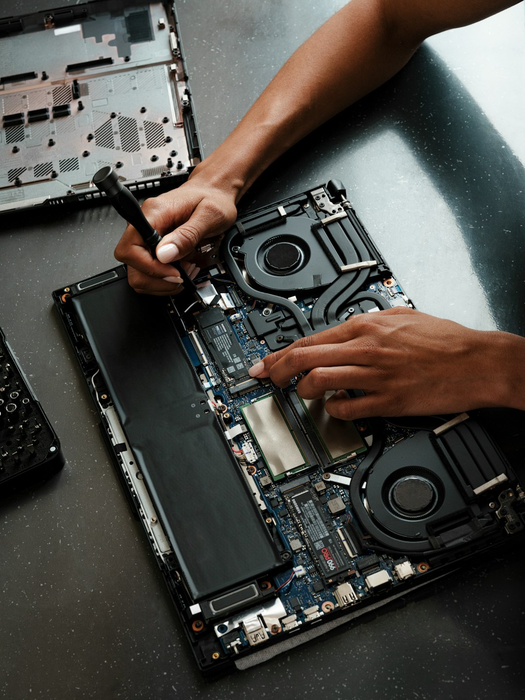 Hands repair a disassembled laptop, using a screwdriver on the motherboard and cooling fans visible inside the chassis.