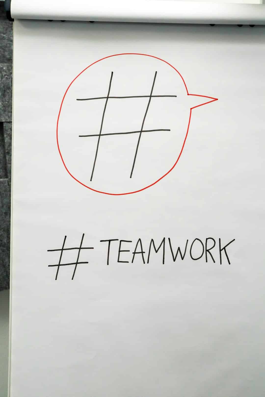 Red outlined speech bubble containing a black tic-tac-toe grid on a whiteboard, with '#TEAMWORK' written below.