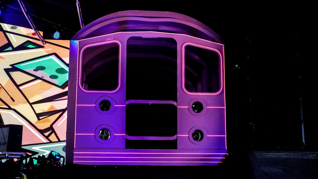 Front of a large pink stage prop shaped like a vintage bus, outlined with neon lights, with circular lights on the sides against a dark stage and a colorful mural background.