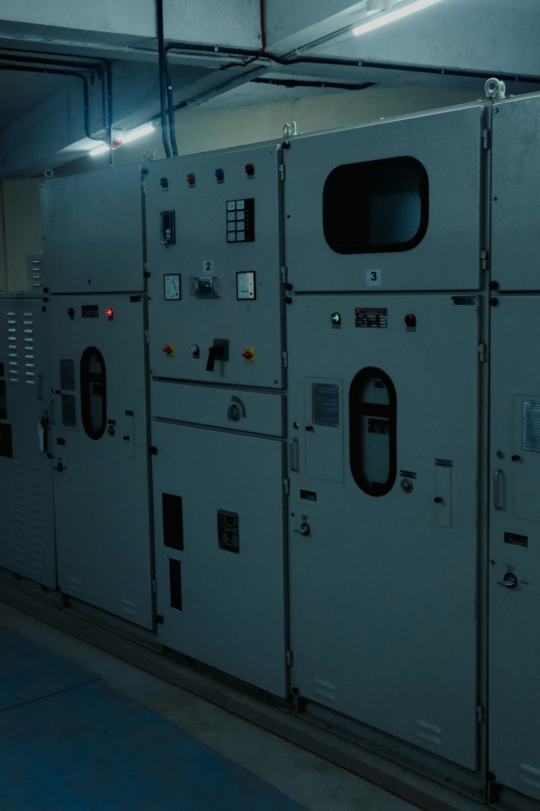 Industrial electrical control panels in a dimly lit utility room with exposed wiring and pipes.