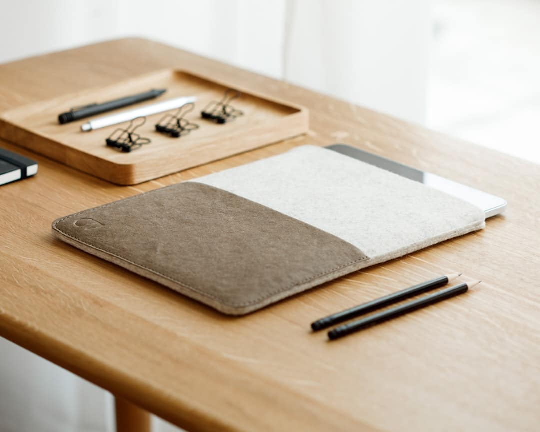 Desk with a two-tone fabric notebook cover resting atop a closed laptop, plus a pencil tray with binder clips and a white pen nearby.