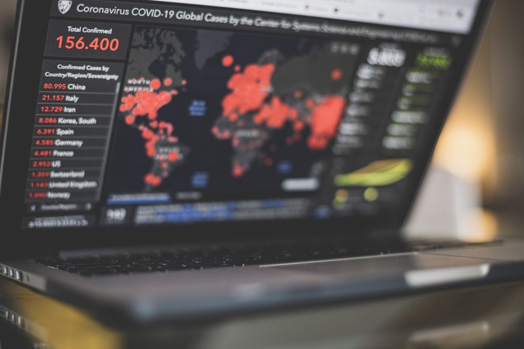 Laptop screen shows a COVID-19 statistics dashboard with a world map marked by red outbreak points and a left sidebar listing country-by-country confirmed cases (e.g., 156,400).