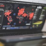 Laptop screen shows a COVID-19 statistics dashboard with a world map marked by red outbreak points and a left sidebar listing country-by-country confirmed cases (e.g., 156,400).