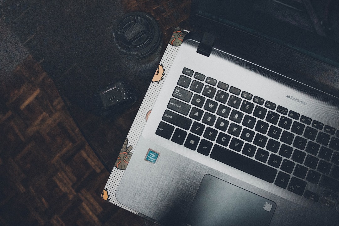 Top-down view of a silver laptop keyboard on a dotted-pattern sleeve, with a camera lens and camera body on a dark wooden desk nearby, viewed from above.