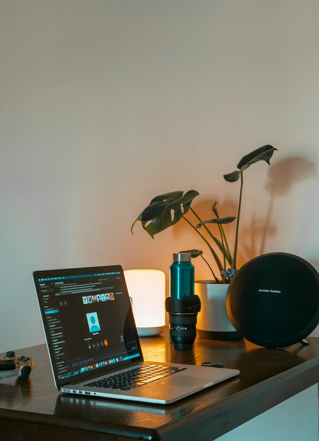 Desk setup: laptop, camera lens, black Harman Kardon speaker, potted plant, lamp, and game controller on a wooden table.