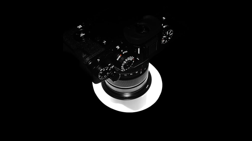 Top-down view of a black camera with a large zoom lens, resting on a circular white highlight against a black background, isolated.