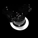 Top-down view of a black camera with a large zoom lens, resting on a circular white highlight against a black background, isolated.