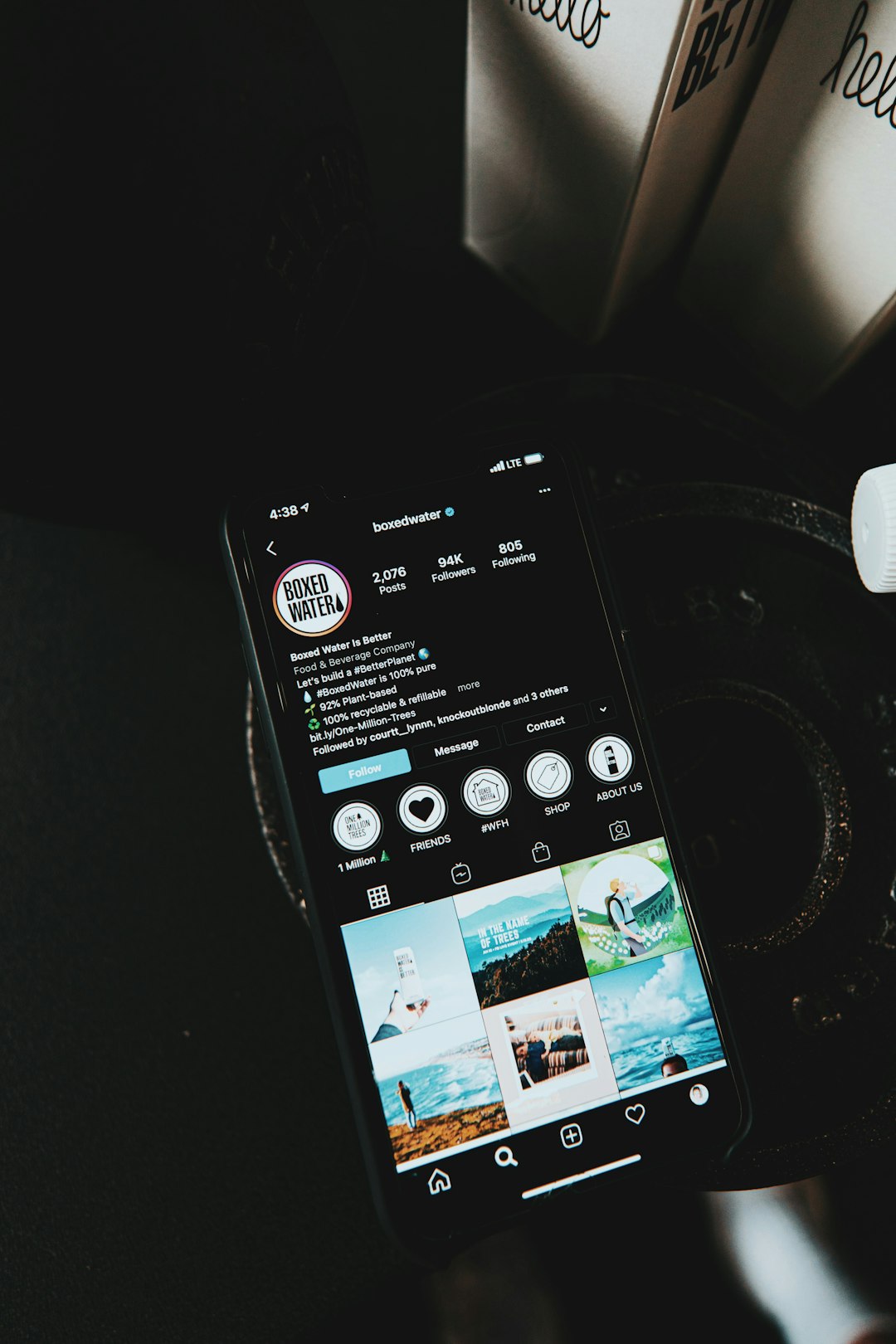 Smartphone on a dark surface showing the Boxed Water Instagram profile with the bio, follower counts, and a grid of photos.