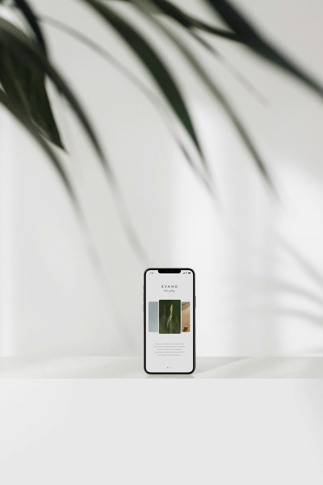 Smartphone on a white desk displaying an app mockup with three image tiles, blurred plant fronds overhead in the background.
