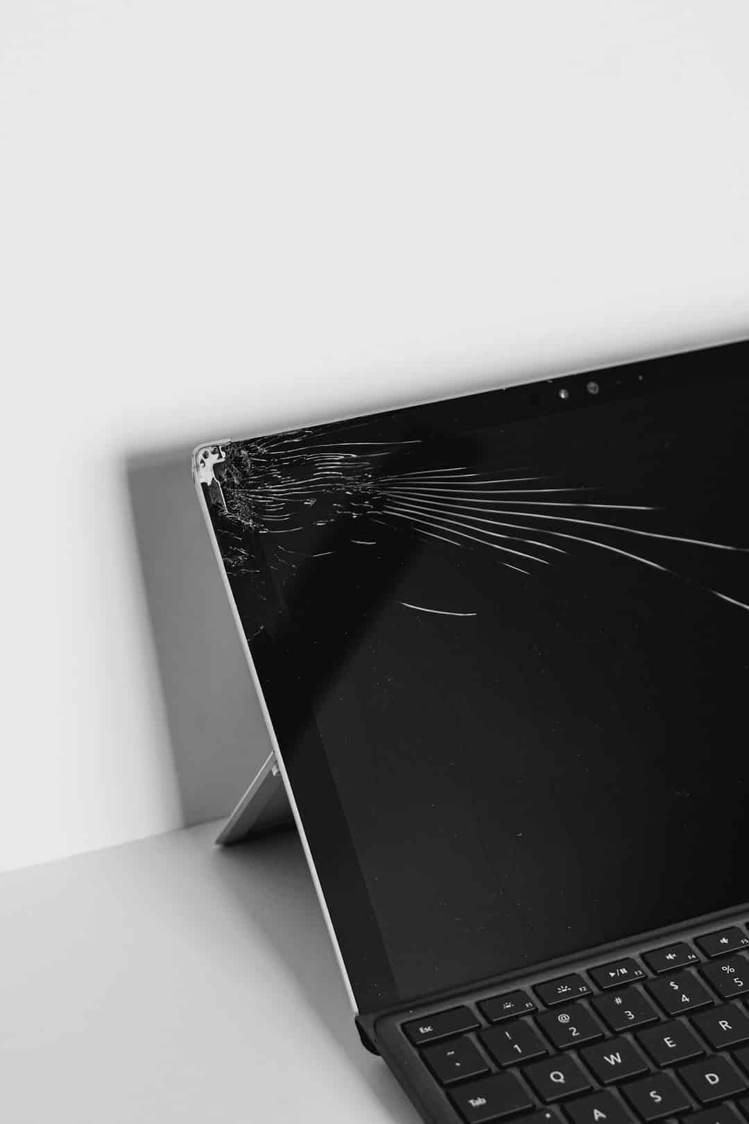 Laptop with a shattered screen near the top-left corner, resting on a stand on a white desk (black-and-white photo).