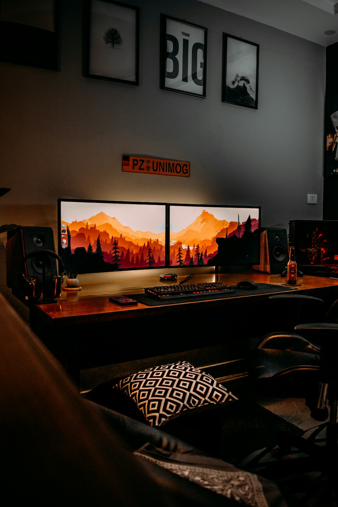 Dual-monitor computer setup on a wooden desk with orange-lit mountain wallpaper, keyboard, mouse, speakers, and a patterned cushion on the floor.