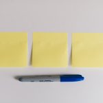 Three yellow sticky notes lined up on a white surface with a blue and gray marker beneath them.