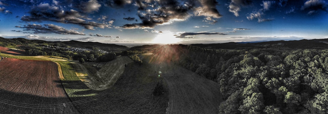 Panoramic sunset over a rural landscape: patchwork fields on the left, dense forest on the right, sun bursting over the horizon.