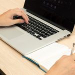 Person typing on a silver laptop while writing in a notebook on a light wood desk.