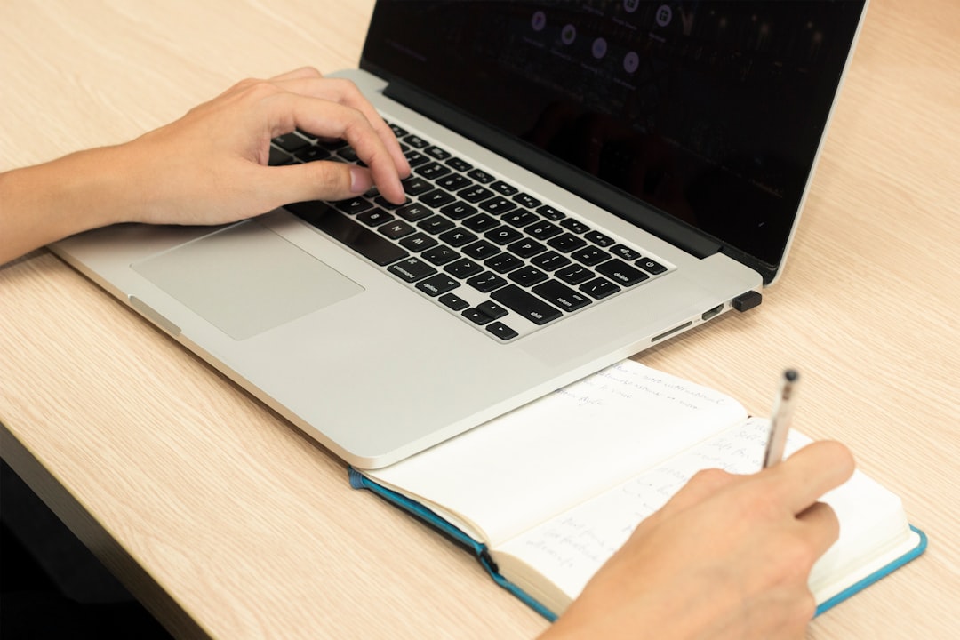 Person typing on a silver laptop while writing in a notebook on a light wood desk.