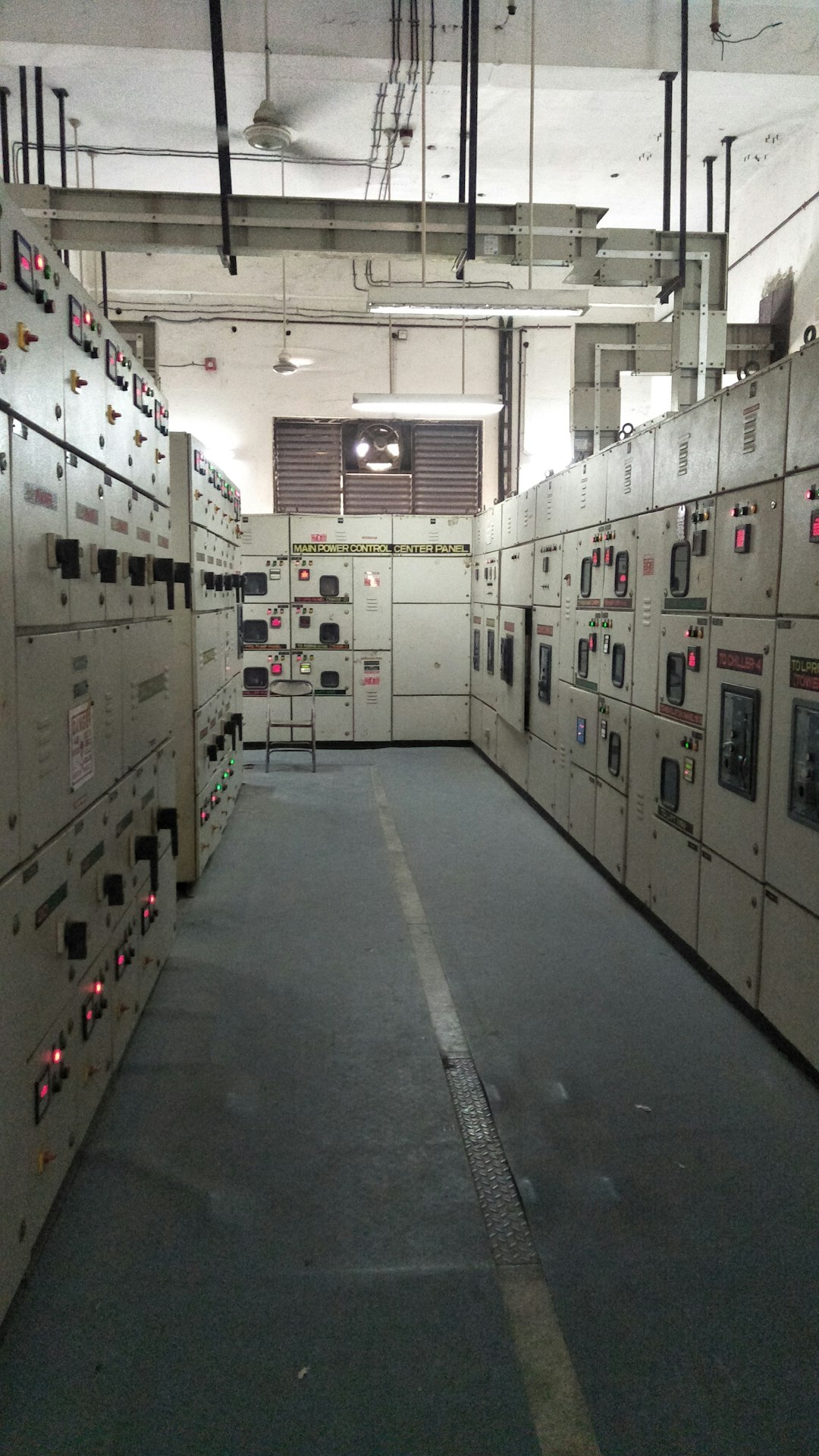 Industrial control room with rows of beige electrical panels, switches and red/green indicator lights on both sides of a narrow aisle.