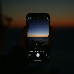 Person holds a smartphone in portrait orientation, capturing a sunset over the ocean; the phone screen shows the camera view with a gradient orange-to-blue sky and silhouettes of buildings along the shore.