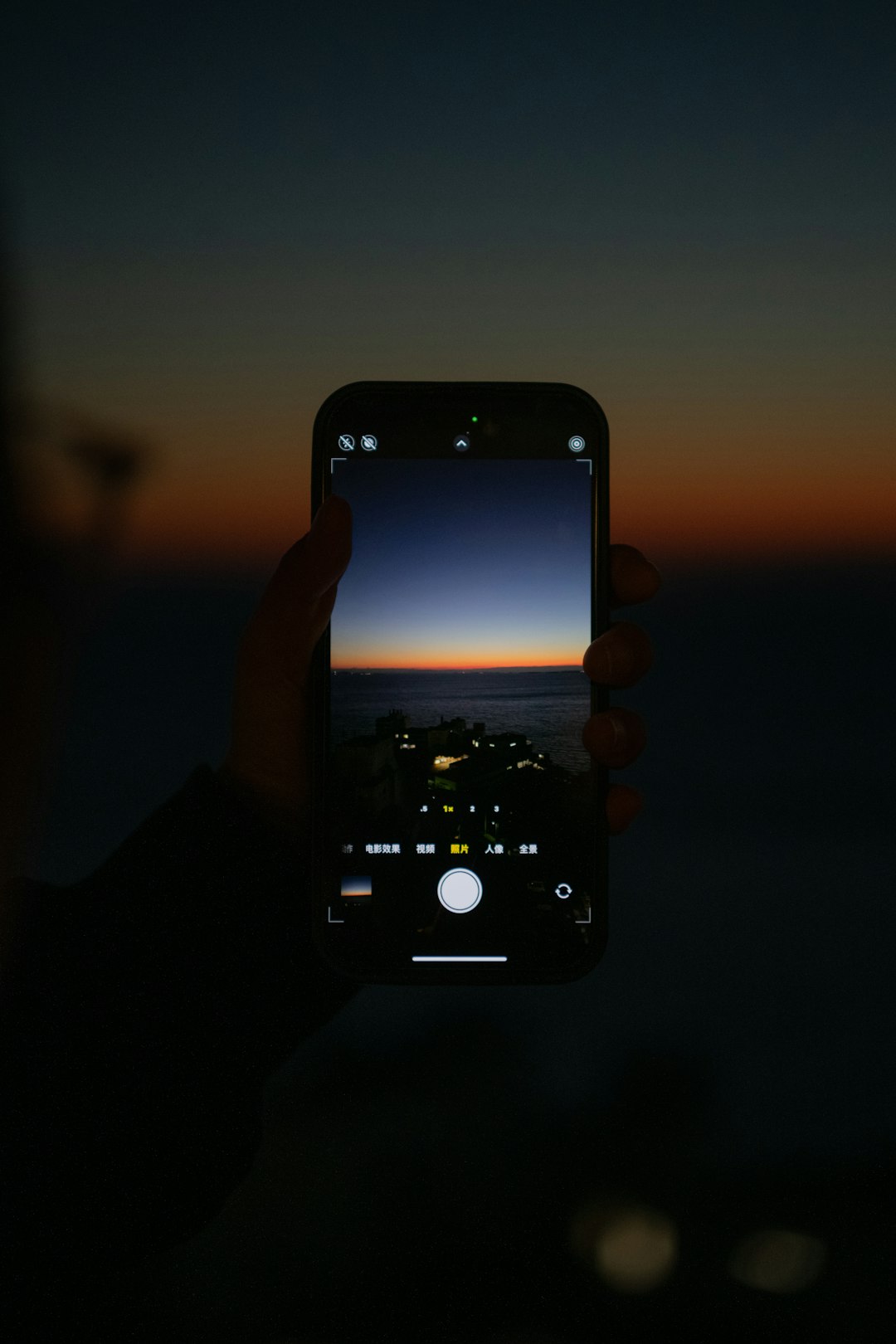 Person holds a smartphone in portrait orientation, capturing a sunset over the ocean; the phone screen shows the camera view with a gradient orange-to-blue sky and silhouettes of buildings along the shore.