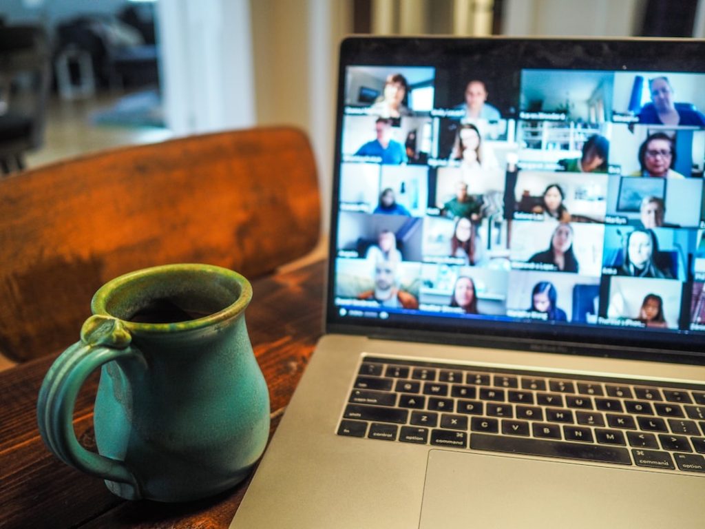 Laptop showing a multi-person video conference on a gallery view; a teal ceramic mug sits on a wooden table beside it.