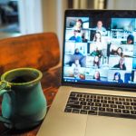 Laptop showing a multi-person video conference on a gallery view; a teal ceramic mug sits on a wooden table beside it.