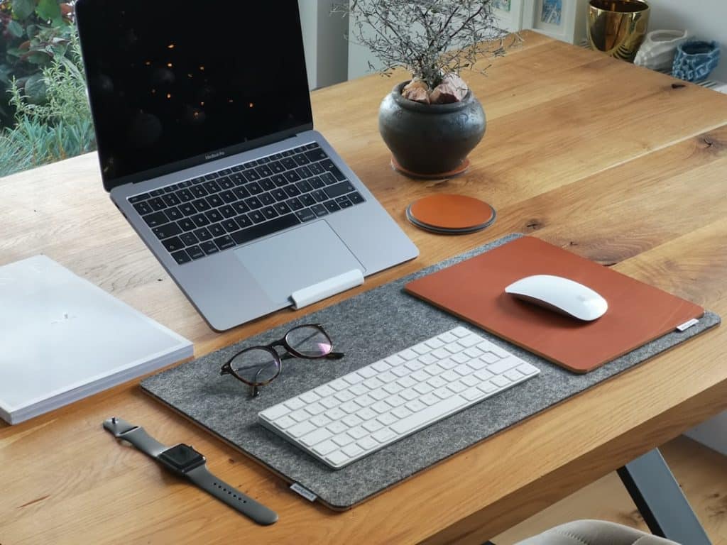 Wooden desk setup with a MacBook Pro, white keyboard and mouse on a felt desk mat, glasses, and a plant in a round pot nearby.