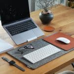 Wooden desk setup with a MacBook Pro, white keyboard and mouse on a felt desk mat, glasses, and a plant in a round pot nearby.