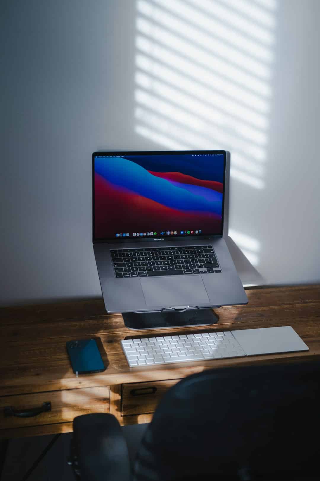 MacBook Pro on a raised stand with a wireless keyboard and a blue smartphone on a wooden desk, sunlit blinds casting stripes.