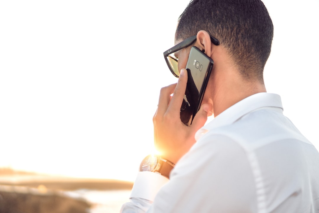 Man in a white shirt talks on a smartphone outdoors at sunset, wearing sunglasses and a wristwatch.