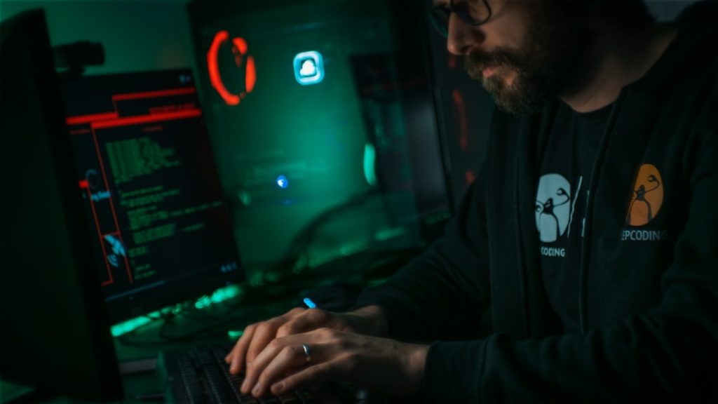 Coder with beard typing on a keyboard in a dimly lit room, green and red neon lights reflecting off monitors and a PC case.