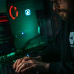 Coder with beard typing on a keyboard in a dimly lit room, green and red neon lights reflecting off monitors and a PC case.