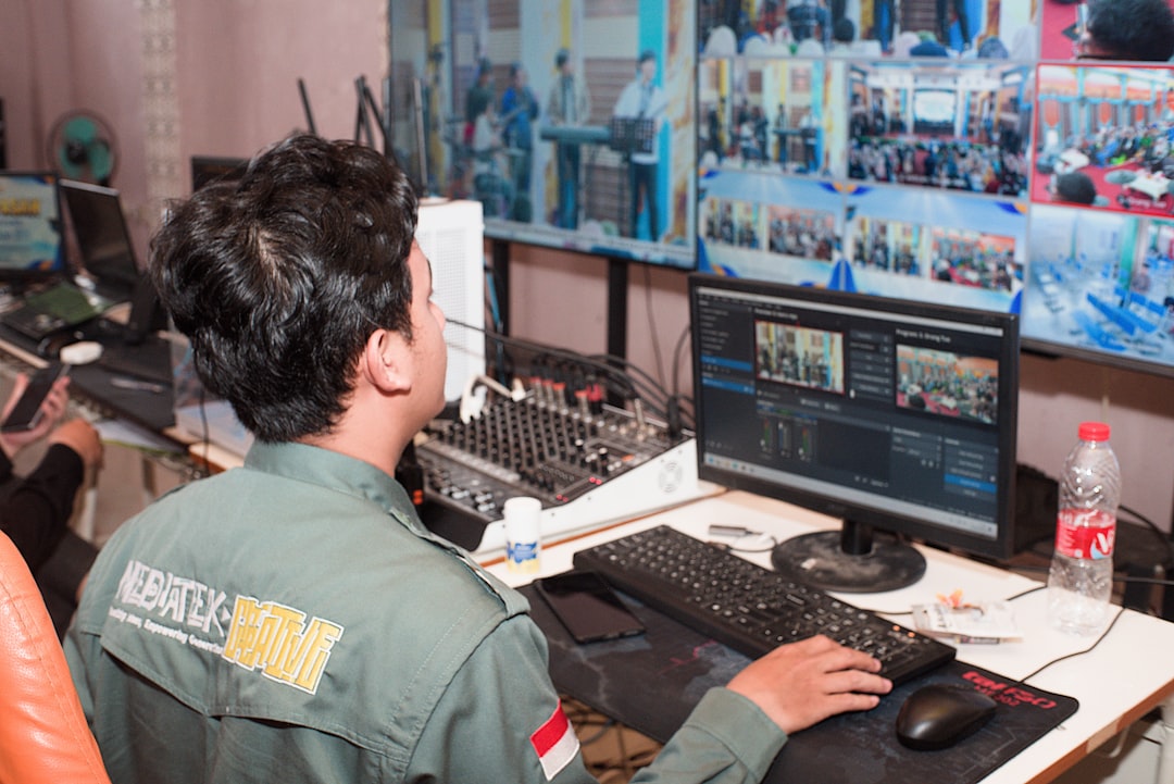 Video editor at a control desk, monitoring and mixing footage on a computer with a wall of clips in the background.